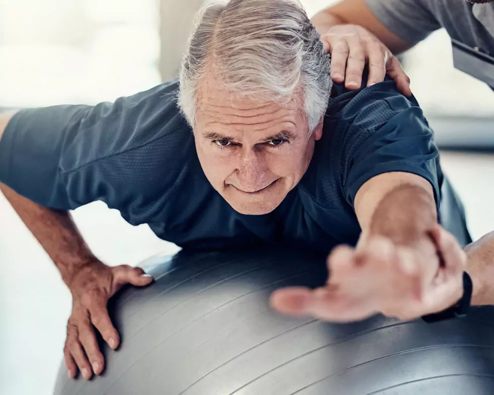 älterer Herr bei Gleichgewichtsübung mit einem Gymnastikball. Physiotherapeut unterstützt und hilft dem Patienten