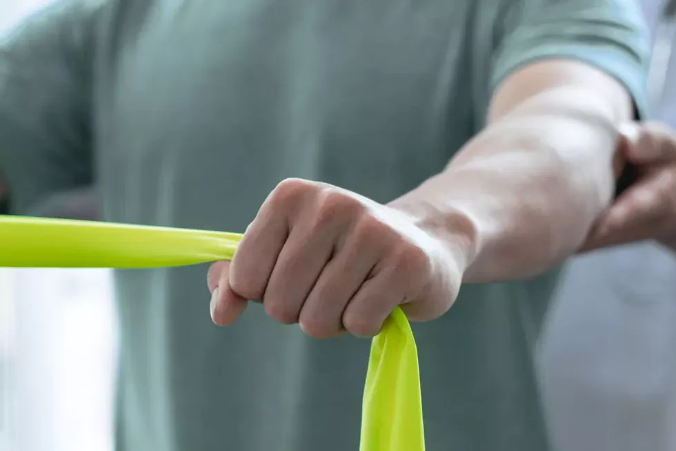 Nahaufnahme eines Patienten, der mit einem Theraband Übung zur Stärkung der Armmuskulatur ausführt. Der Physiotherapeut hält unterstützend die Arme des Patienten