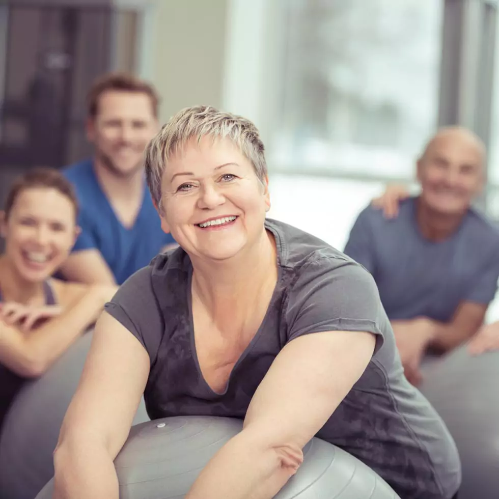 Lächelnde ältere Frau, die einen Pilates-Kurs im Fitnessstudio genießt und sich auf ihren Ball stützt und in die Kamera lächelt, während die Klasse hinter ihr steht