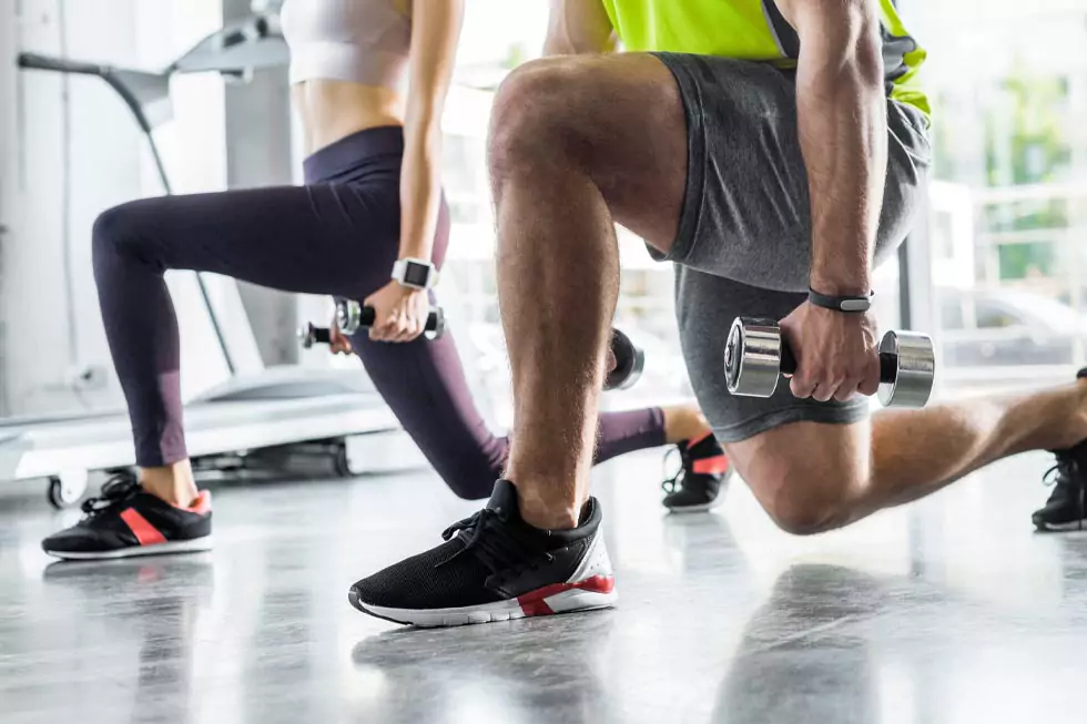 Ausschnitt eines Sportlers und einer Sportlerin bei Ausfallschritten mit Hanteln in einem Sportzentrum