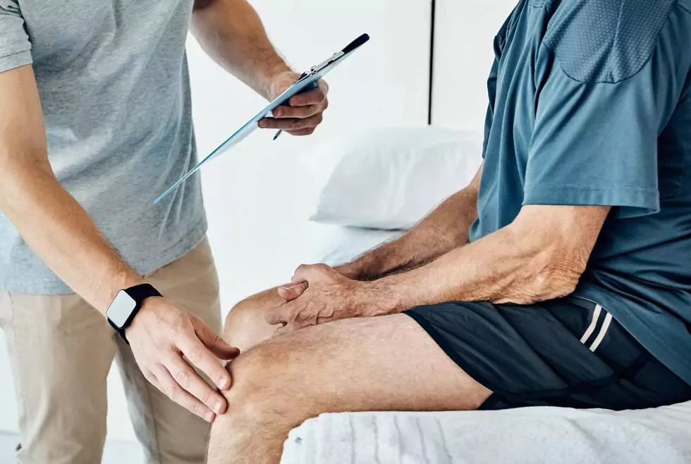 Physiotherapeut untersucht das Knie eines älteren Patienten. Physiotherapeut hält die Patientenakte in der Hand