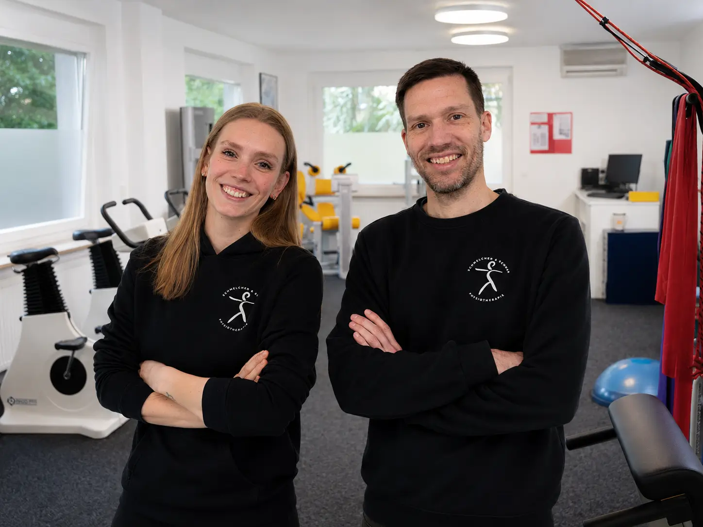 Zwei Personen mit verschränkten Armen tragen schwarze Pullover mit weißem Logo in einem Fitnessraum mit Trainingsgeräten